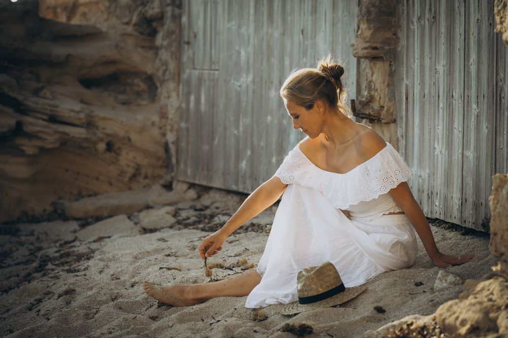 Farbfoto einer barfüßigen Frau in weißem schulterfreiem Kleid am Strand auf Mallorca, die mit einem Stock den Sand berührt.