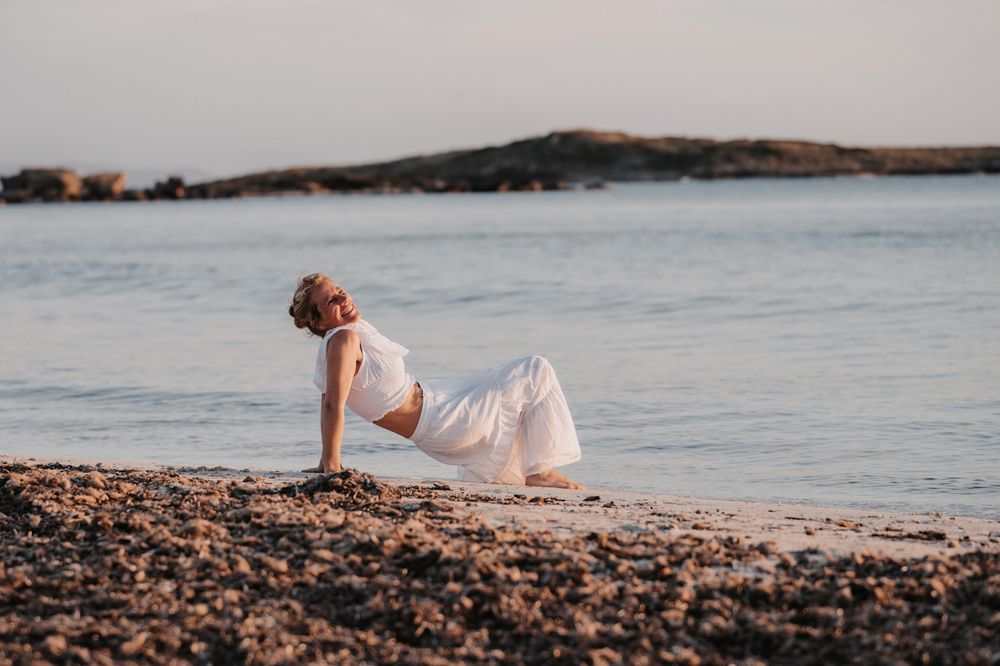 Farbfoto einer Frau in weißer Kleidung am Strand, in Rückbeuge-Brücke, lachend und zum Himmel blickend.
