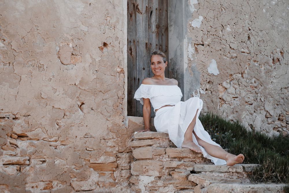 Farbfoto einer Frau in weißer Sommerkleidung, barfuß auf Stufen eines verfallenen Gebäudes auf Mallorca sitzend.