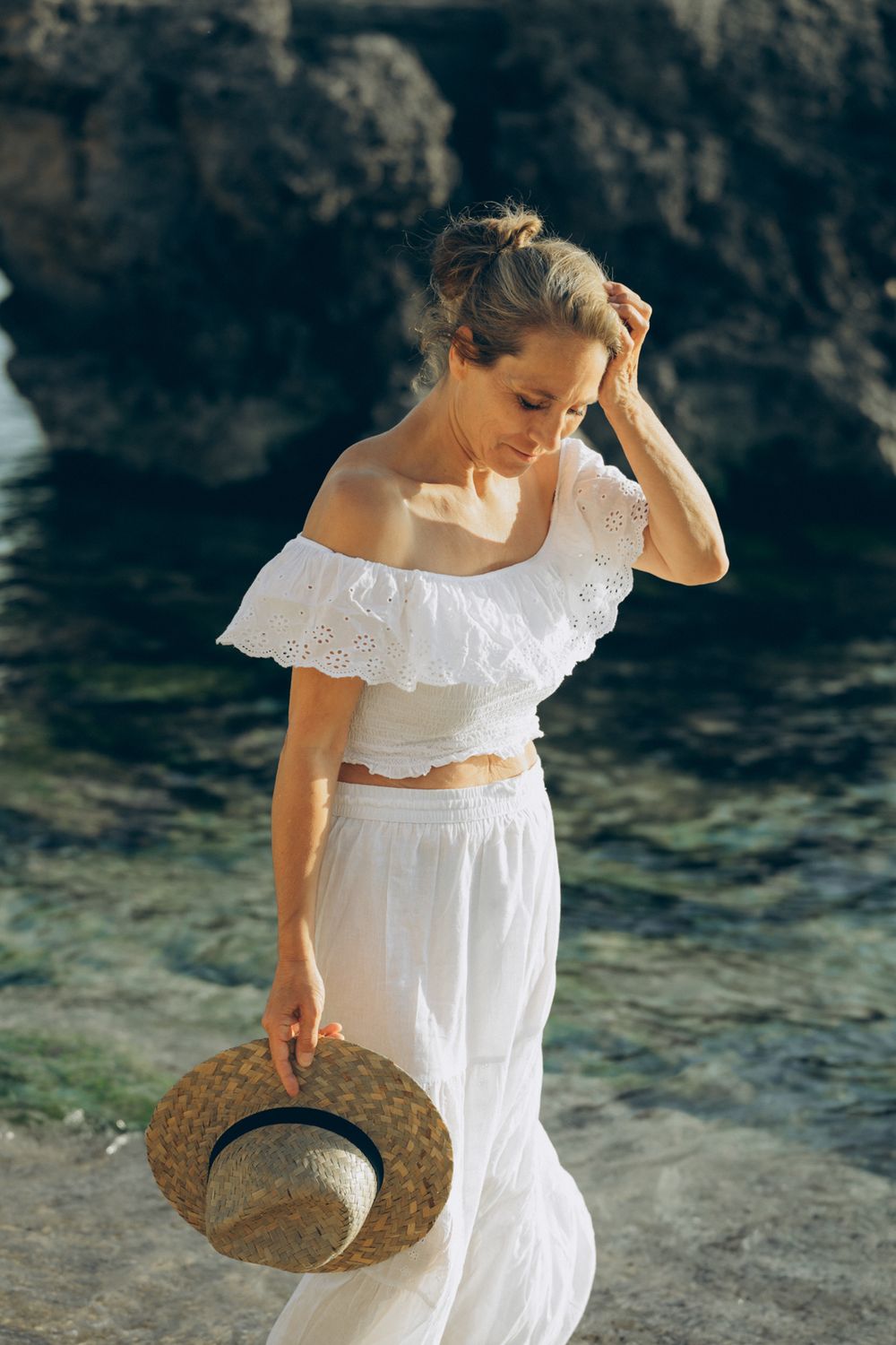 Farbfoto einer Frau in weißer Leinenkleidung im Wasser, auf Mallorca, wischt sich eine Strähne aus dem Gesicht, Hut in der Hand.