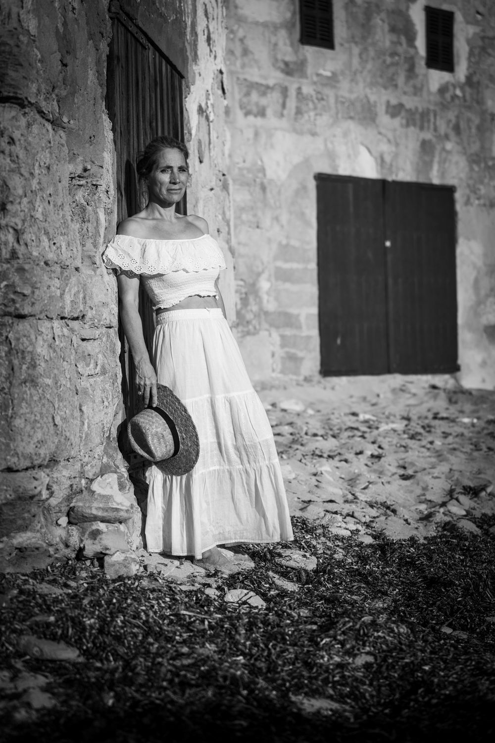 Schwarz-Weiß-Foto einer Frau in weißem Rock und Carmenbluse, Hut in der Hand, an einem steinernen Gebäude lehnend.