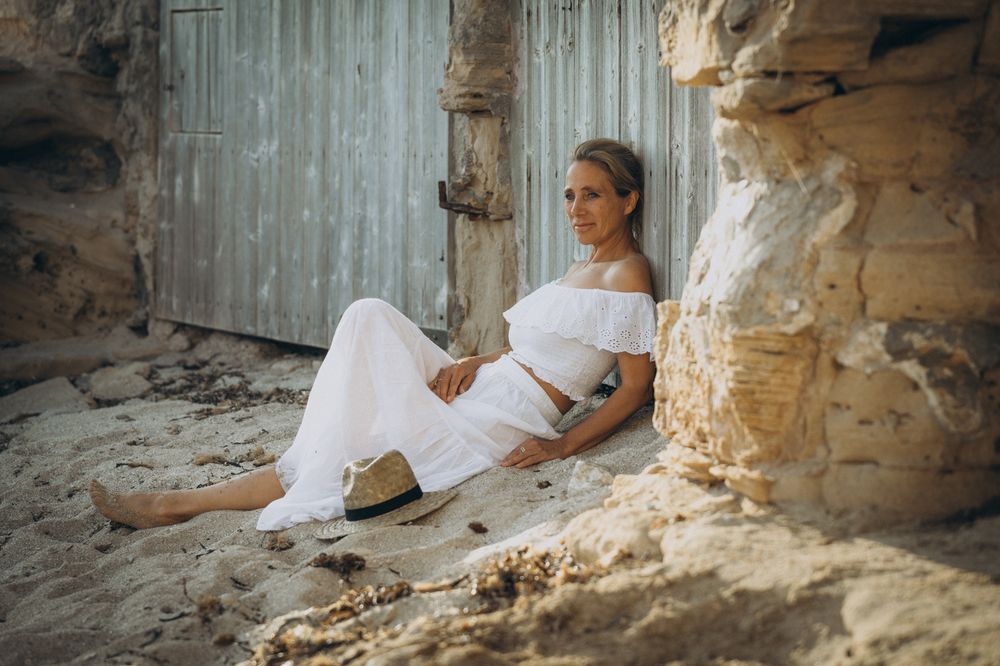 Farbfoto einer Frau in weißer Sommerkleidung, am Strand auf Mallorca an ein Holztor gelehnt, ruhige Stimmung.
