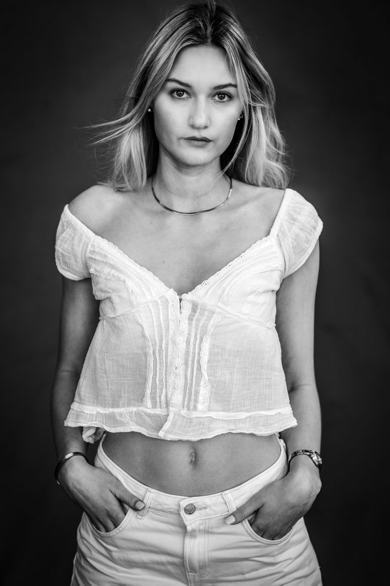 Schwarz-Weiß-Portrait einer jungen Frau in Weiß mit Händen in den Hosentaschen, angeschnittenem Kopf und ernstem Blick in die Kamera.
