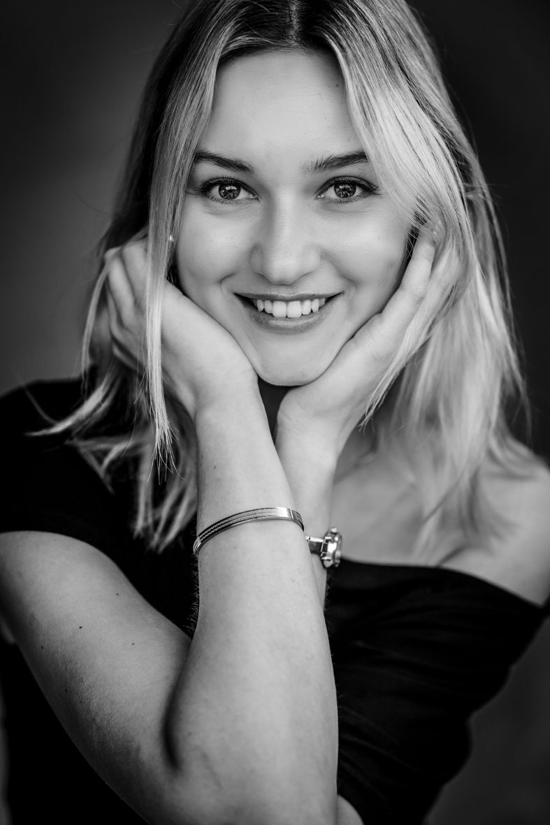 Schwarz-Weiß-Portrait einer jungen Frau im schwarzen Oberteil, die ihren Kopf auf die Hände stützt und herzlich in die Kamera lacht.