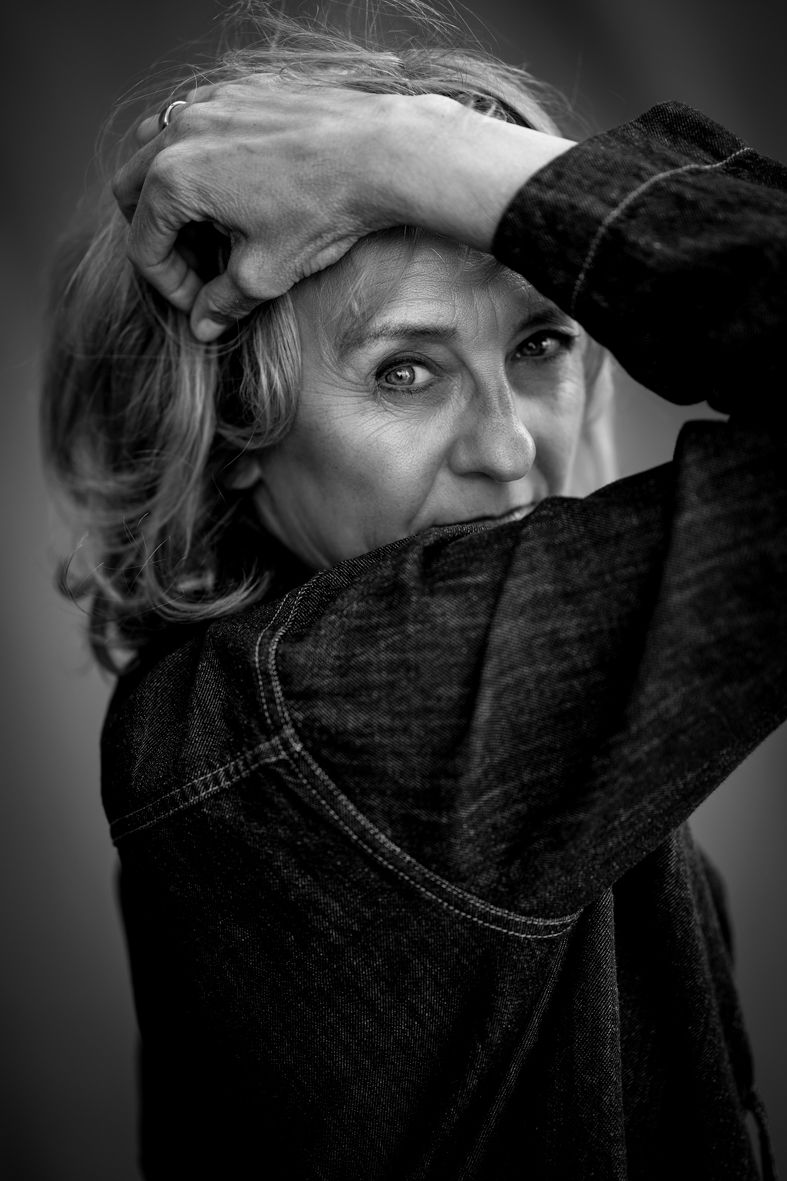 Schwarz-Weiß-Portrait einer Frau im Jeanshemd, die den Arm ans Gesicht hält und mit intensivem Blick durch den Arm in die Kamera schaut
