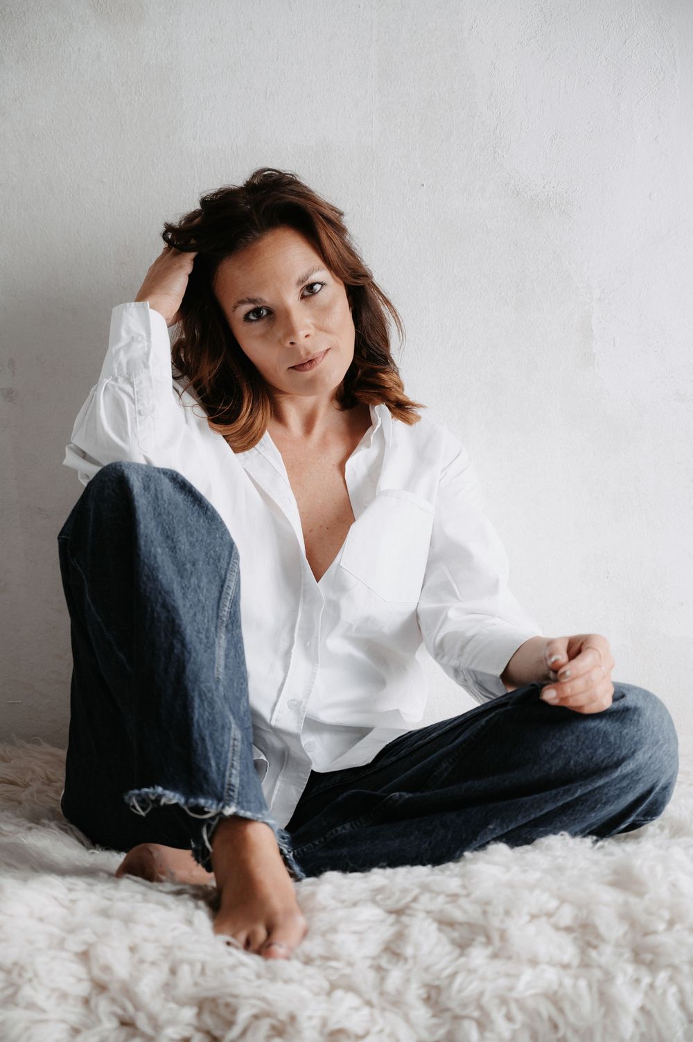 Frau in Jeans und weißer Bluse im Studio, stützt sich mit dem Ellbogen aufs Knie und blickt in die Kamera – natürliches Licht, klarer Stil.