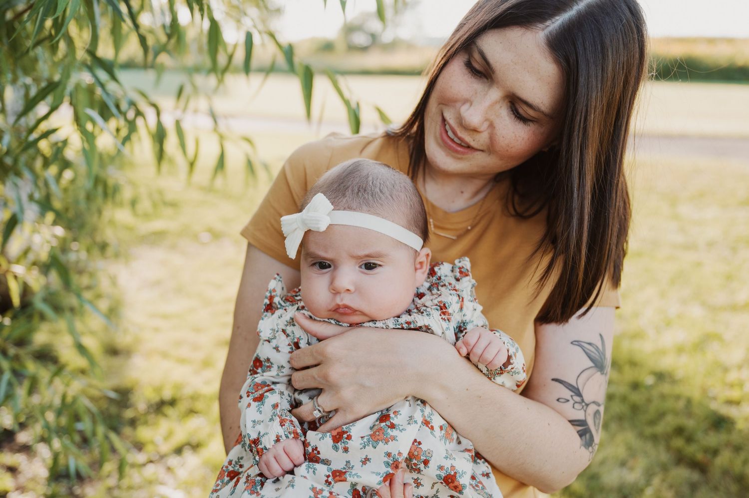 A loving parent holds a baby wearing a floral dress and white bow headband in a sunny outdoor setting with willow trees.