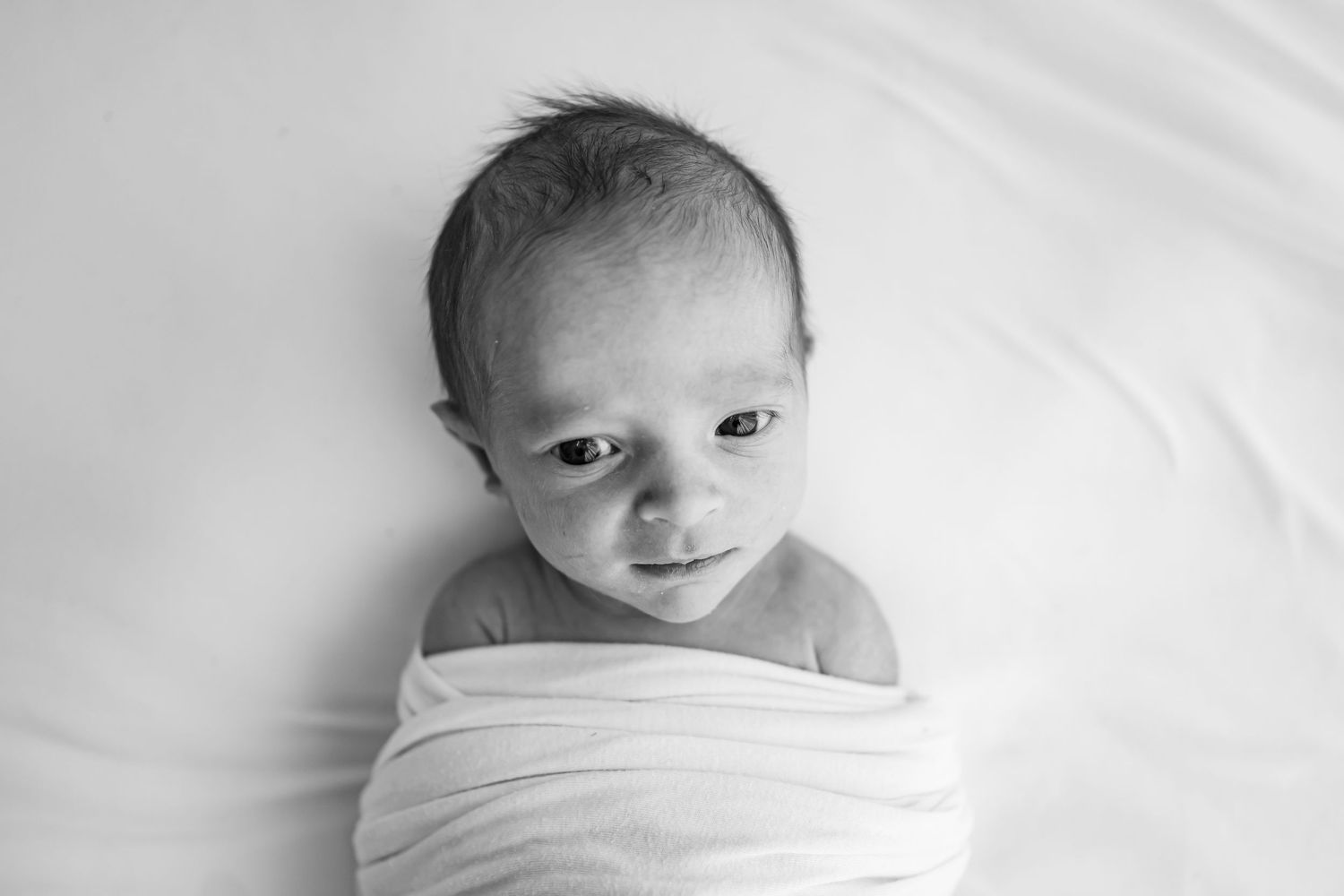 Black and white photo of a tiny baby swaddled in white cloth looking up at the camera against white background.