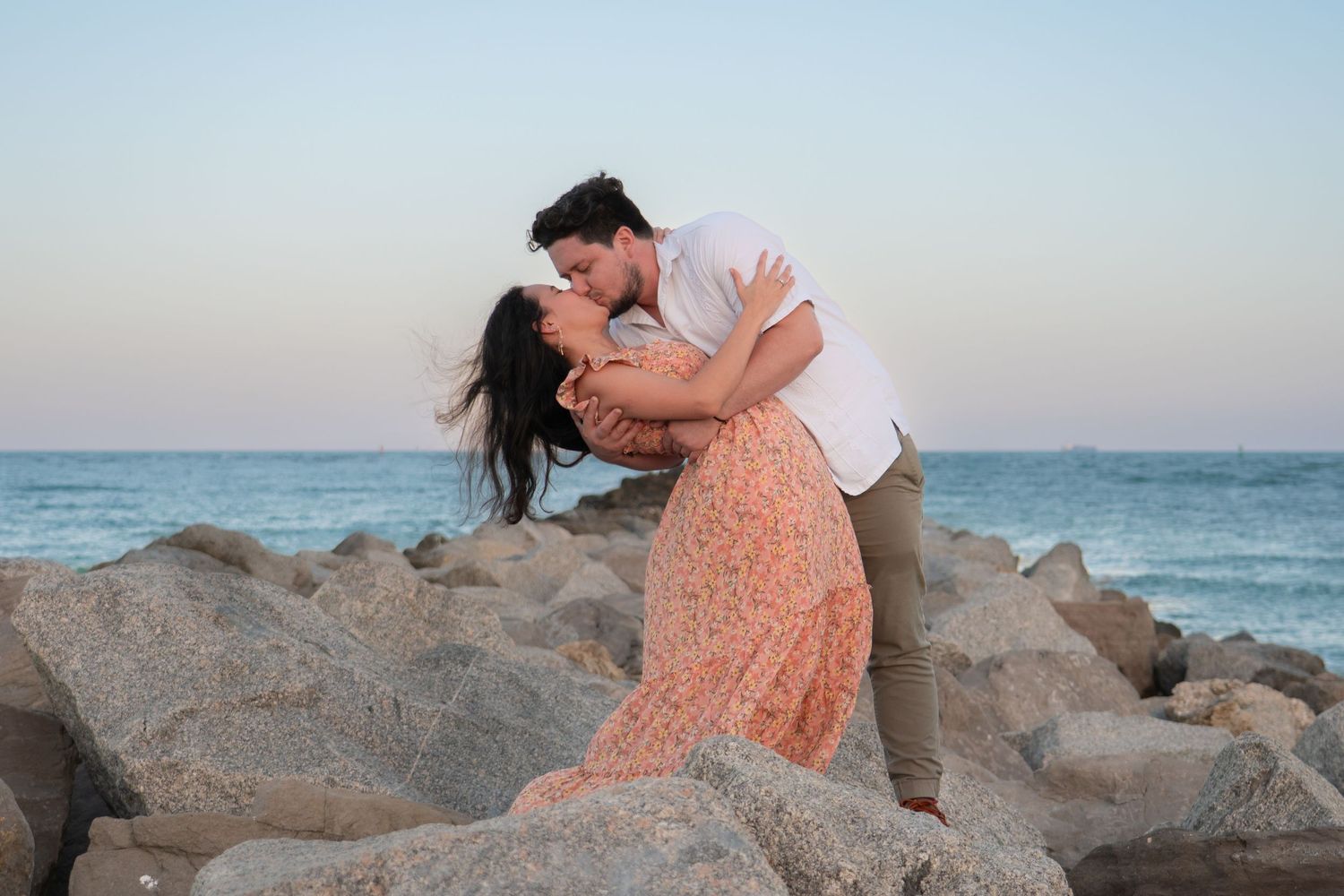 Romantic couple sharing a passionate kiss on rocky shoreline at sunset by the ocean.