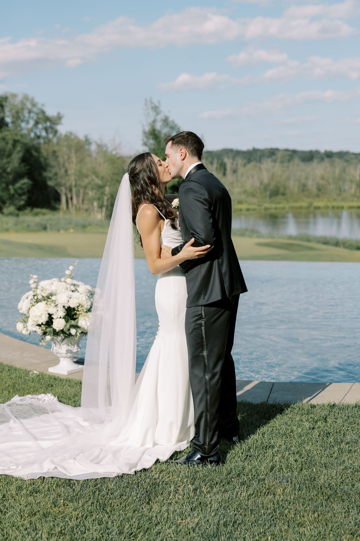 A bride and groom share a romantic kiss by a serene lake, with white floral arrangements and a flowing veil in view.