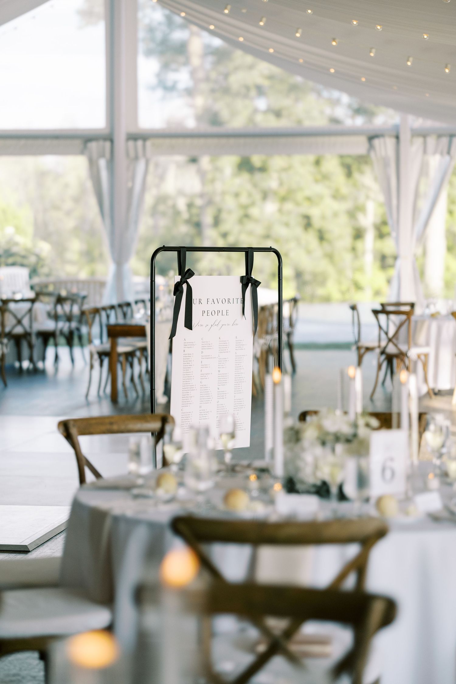 Elegant wedding reception setup in white tent with string lights and cross-back chairs illuminated by natural light.