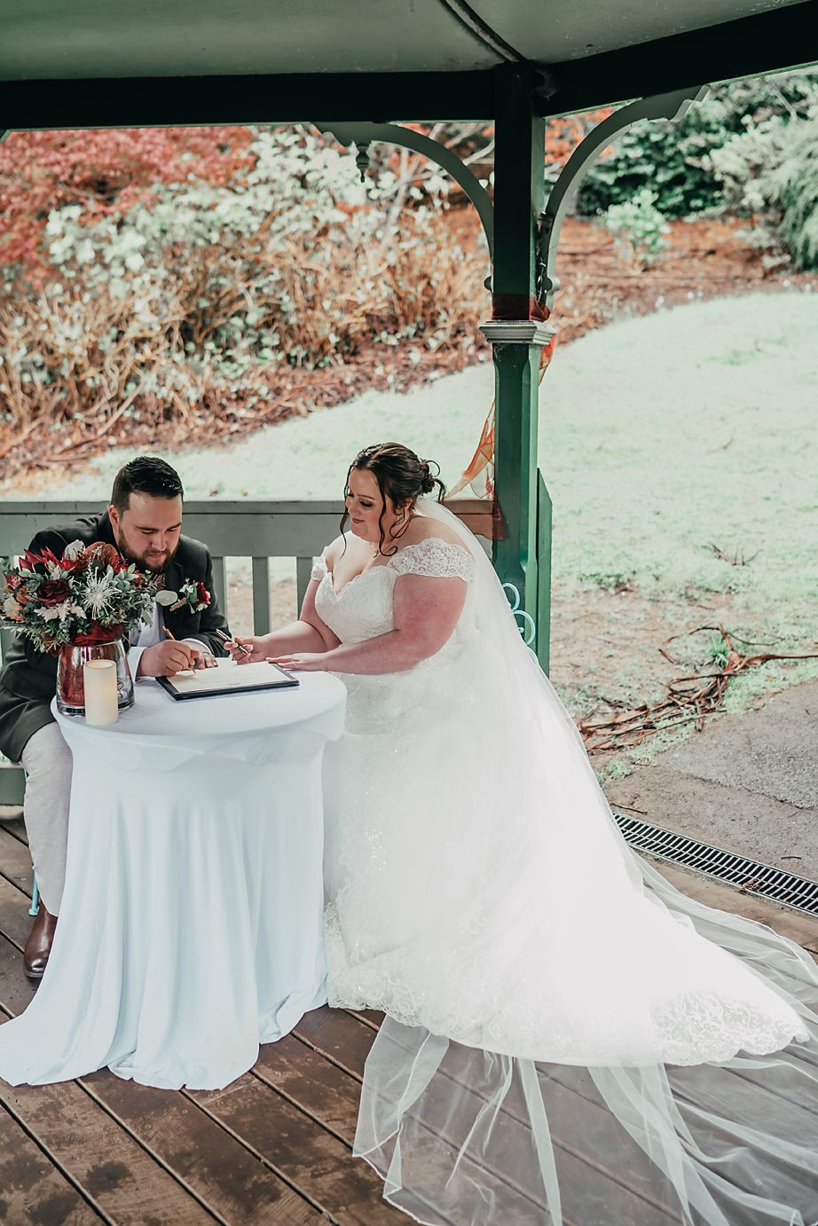 Two people in elegant wedding attire sit at a round table covered in white cloth during an outdoor reception.