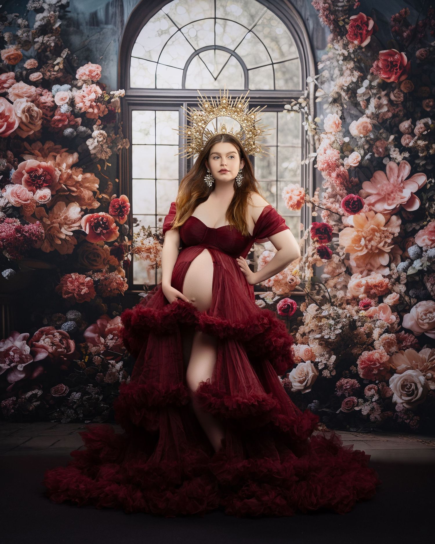 A dramatic fashion scene with a flowing burgundy gown and gold crown against a moody floral backdrop by a window.