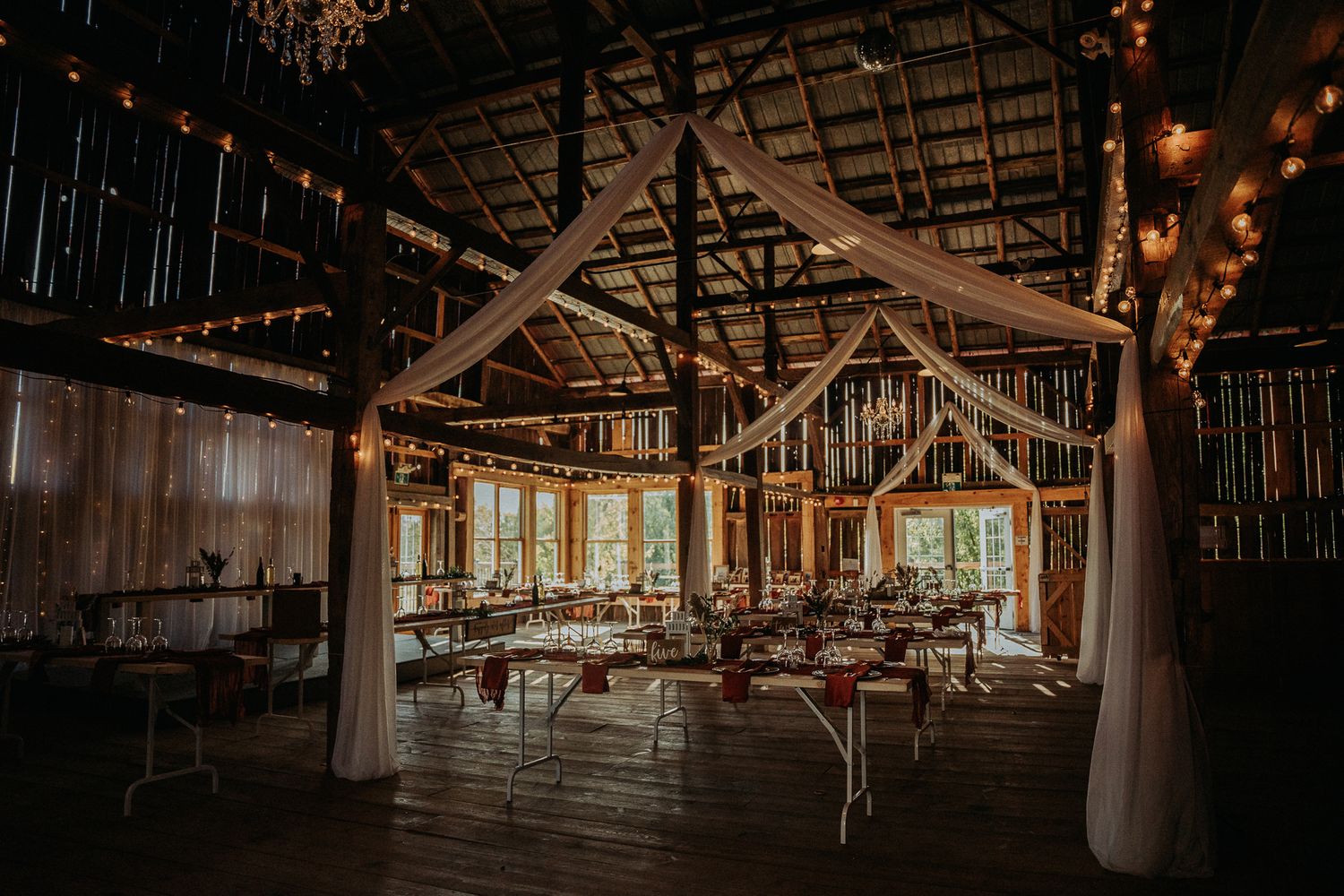 Rustic barn wedding reception venue with white draping and string lights illuminating wooden tables and chairs.