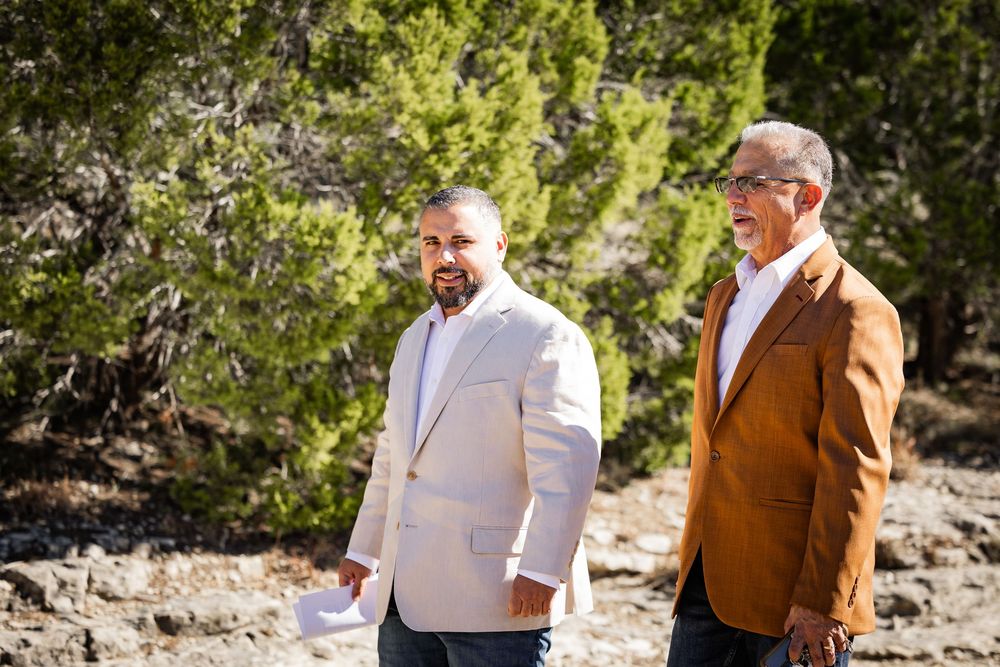 Two men in blazers stand together outdoors on a sunny day with green foliage in the background.