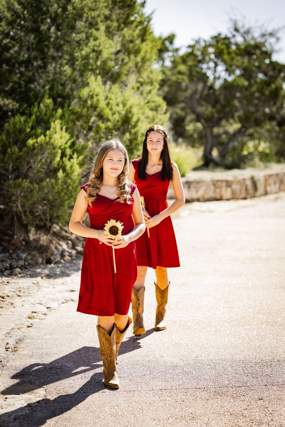 Two figures in red dresses and brown boots walk along a sunny path holding sunflowers.