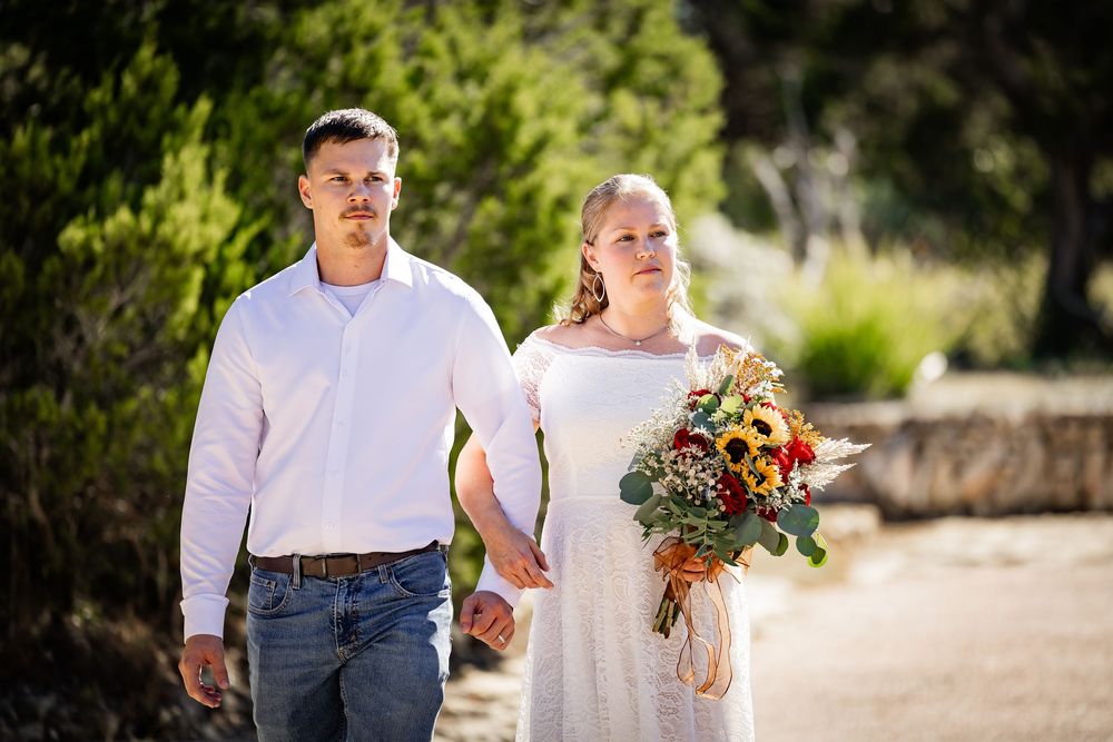 A couple walks outdoors in a sunny garden setting with colorful flowers and greenery.