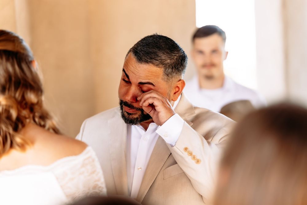 Emotional groom wipes away tears during wedding ceremony while wearing a light colored suit and bowtie.