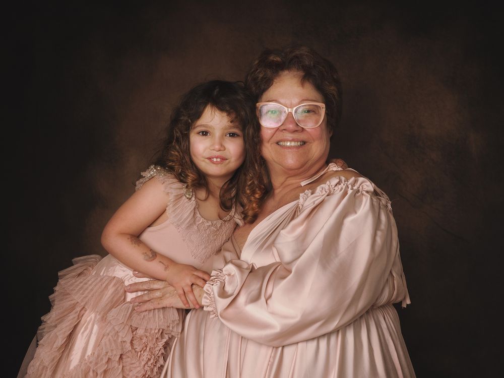 A heartwarming professional portrait shows two generations wearing matching soft pink formal dresses against a dark backdrop.