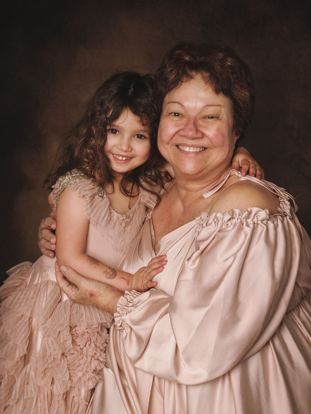 A vintage portrait showing a warm embrace between an older person in pink satin and a young child in a dusty rose dress.