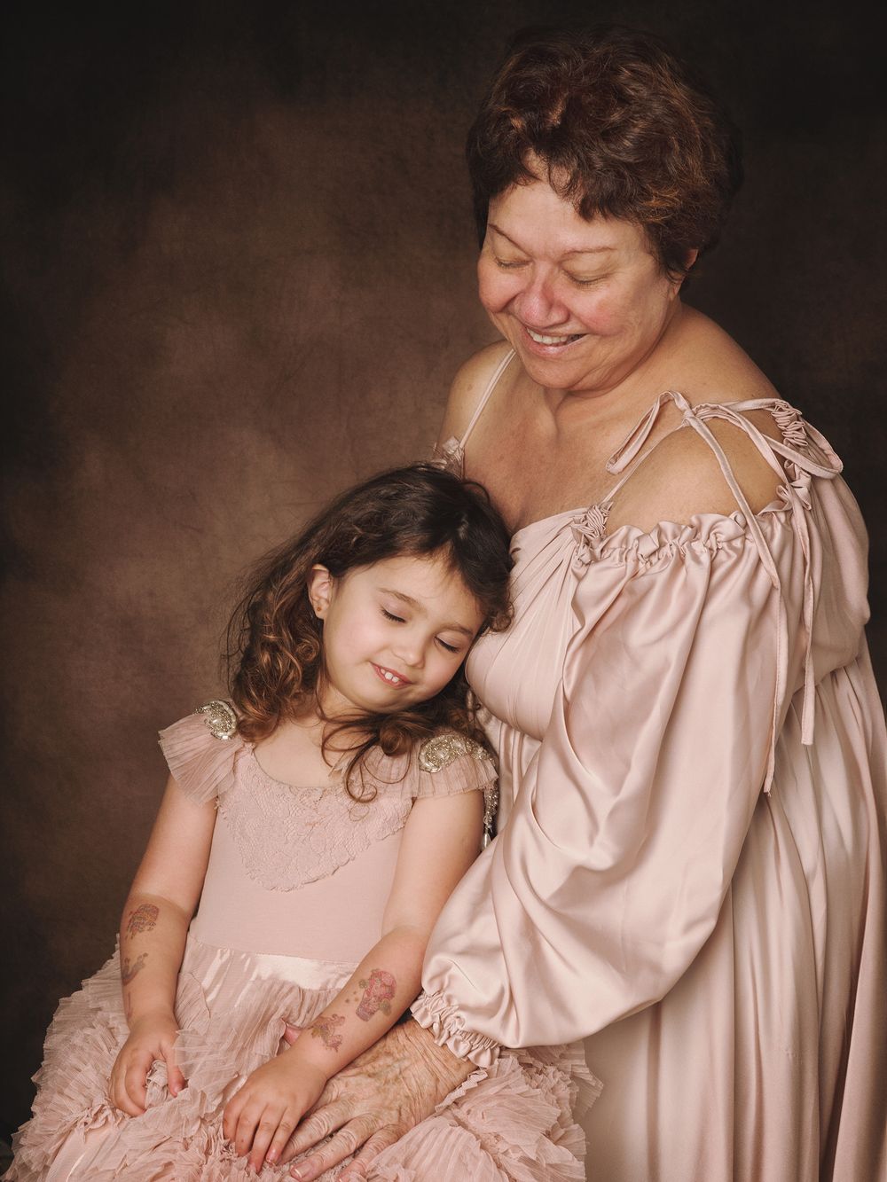 A tender portrait showing joyful smiles and shared affection between two figures in matching pink dresses against brown backdrop.