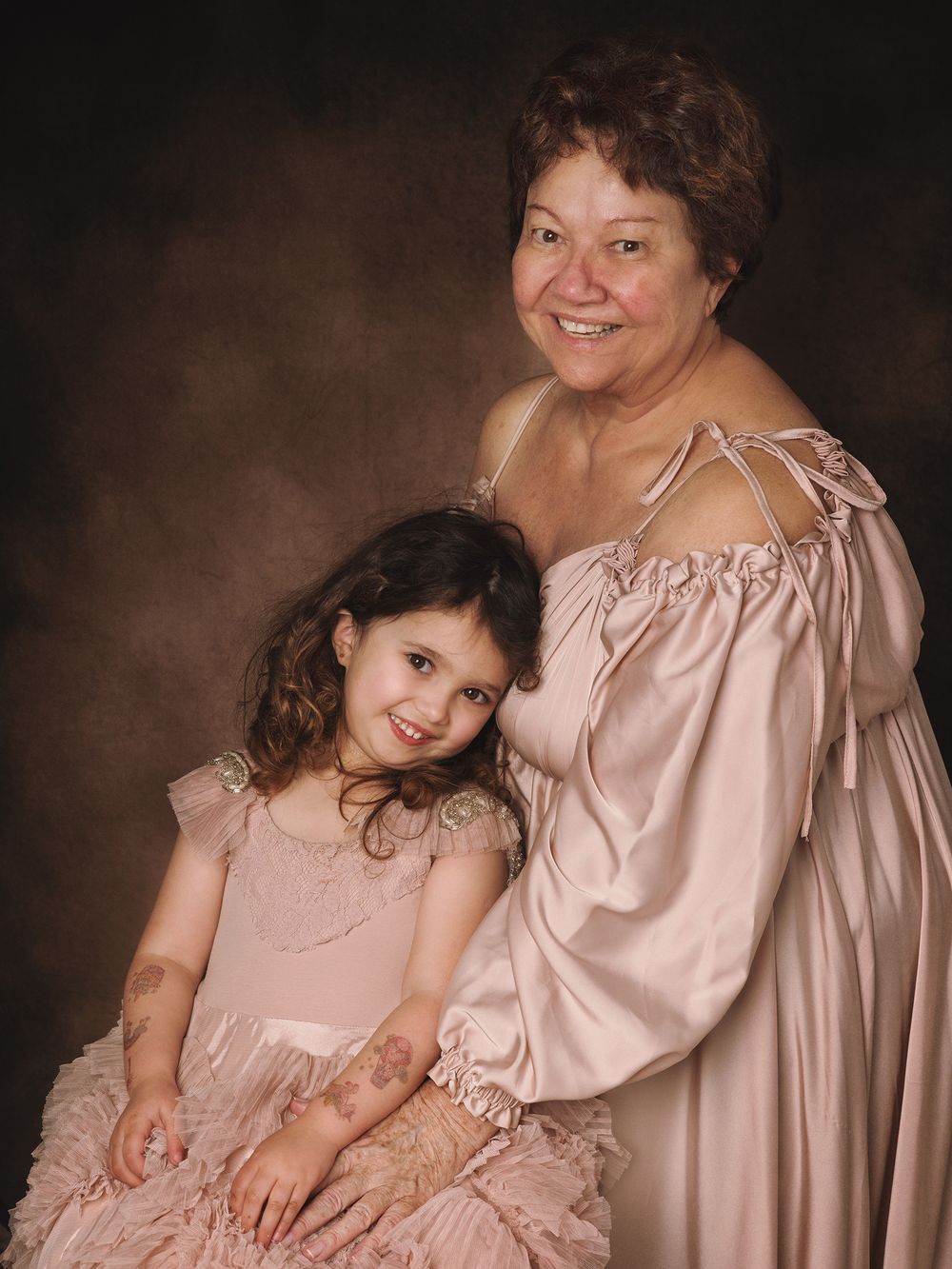 A heartwarming studio portrait shows two people in pink and beige dresses posed together against a dark brown background.