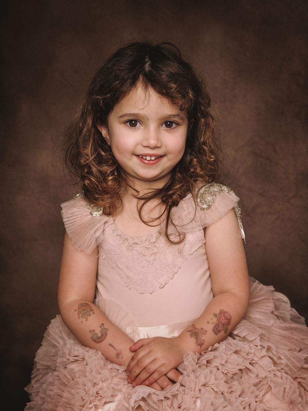 A young child in a pink lace dress poses for a formal portrait against a dark brown backdrop.