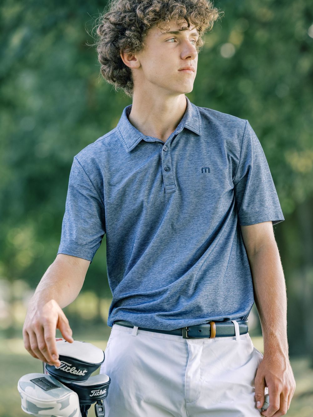 A golfer wearing a blue polo shirt and white pants stands outdoors holding a Titleist golf club against a green background.