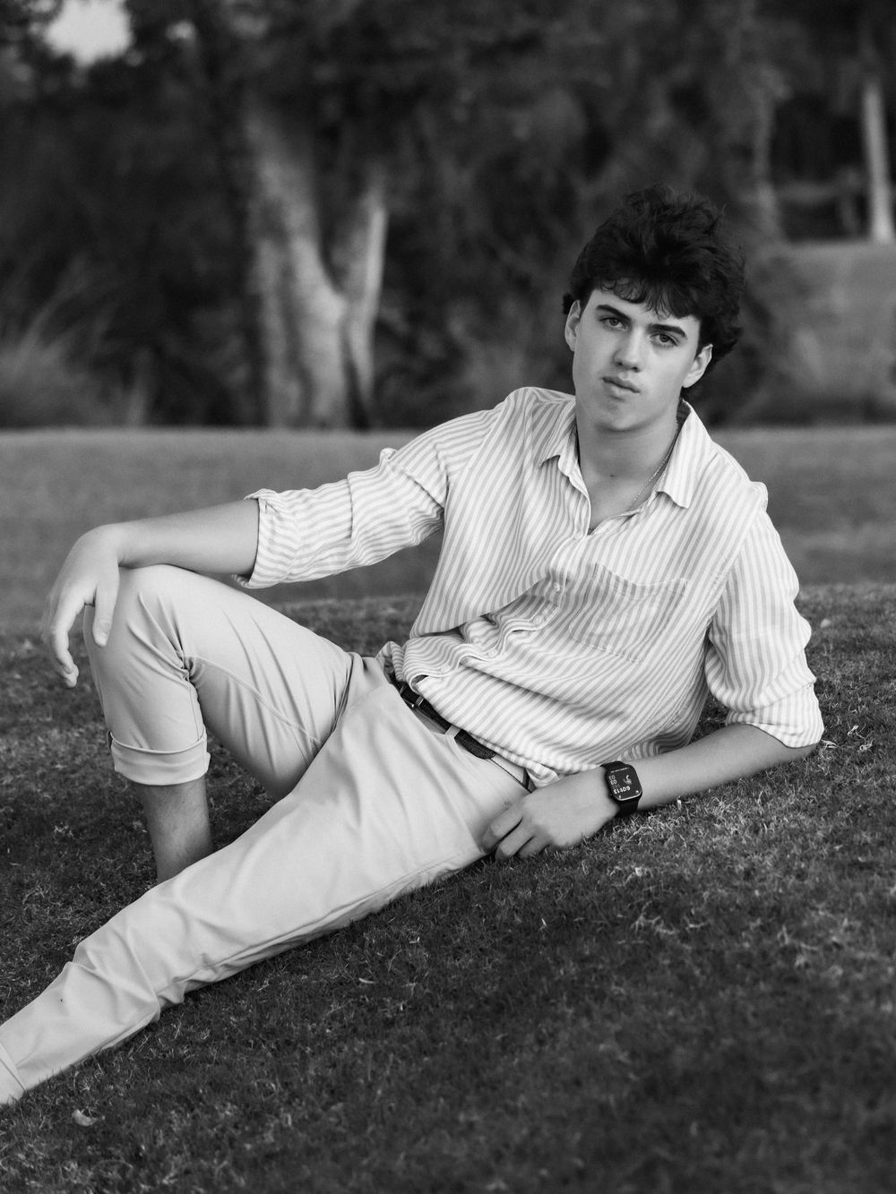 Black and white portrait of someone in a striped button-down shirt and light pants lounging on grass outdoors.