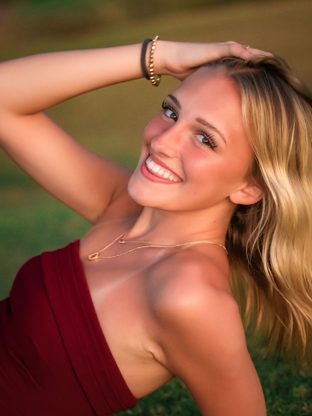 A person in a burgundy top poses outdoors with a bright smile and blonde hair while wearing delicate jewelry.