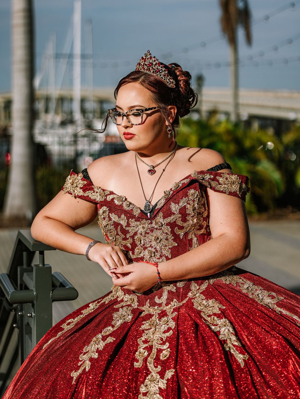 A person in a stunning red and gold quinceañera dress with glasses poses outdoors near a marina.