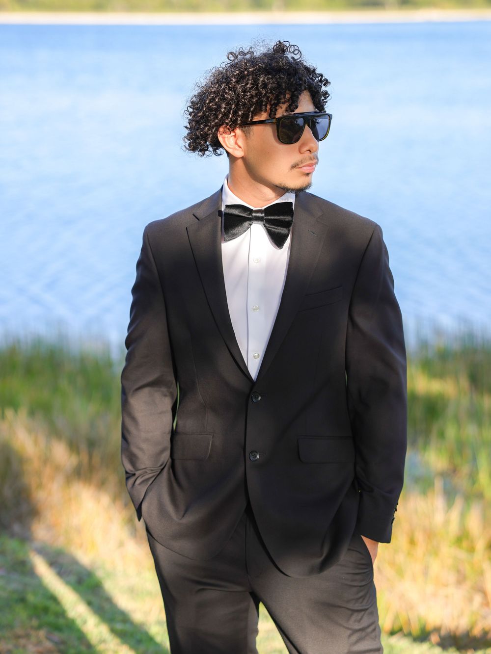A well-dressed figure in a formal black tuxedo and bowtie with sunglasses poses outdoors by a lake.