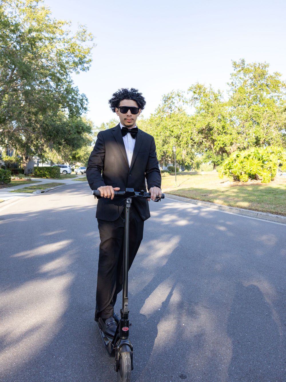 A person riding an electric scooter down a sunny suburban street while wearing a formal black suit and sunglasses.