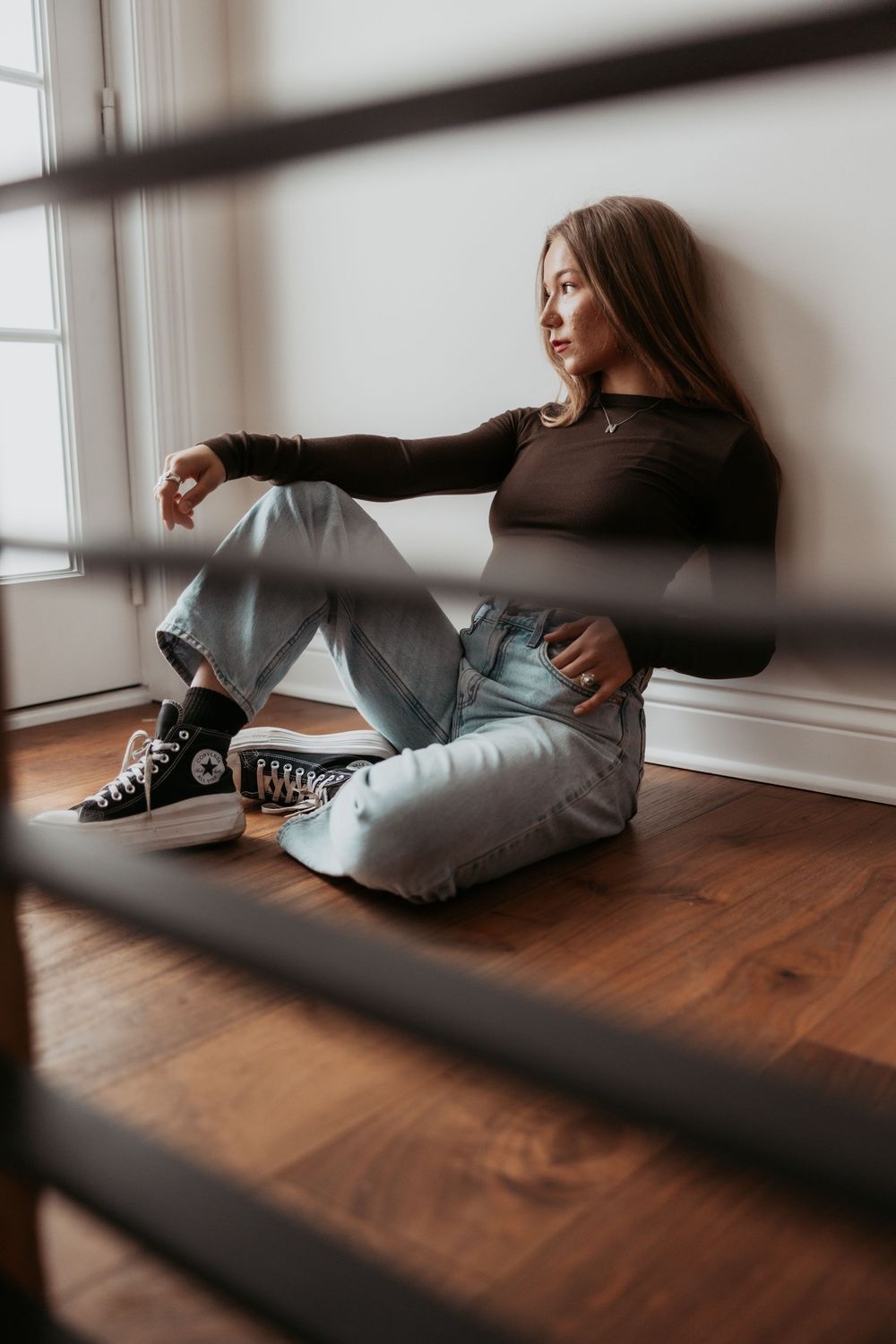 Moody lifestyle portrait of someone sitting on hardwood floor by window wearing black sweater, jeans and high-top sneakers.