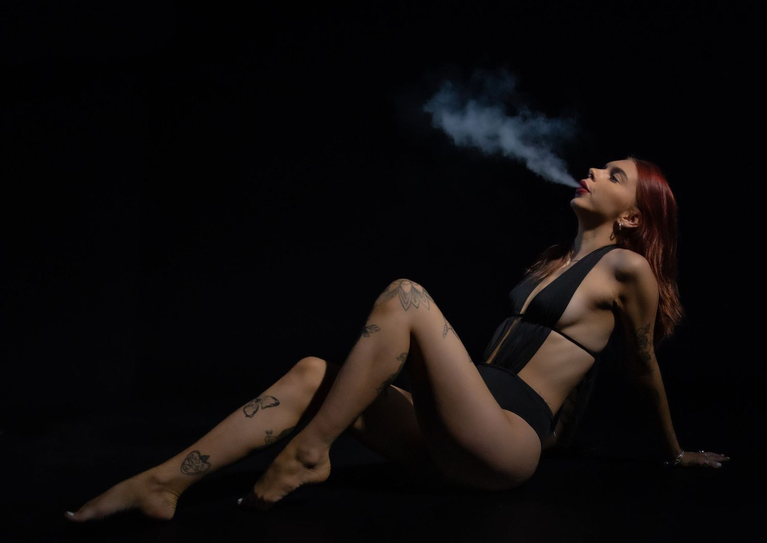 Artistic dark portrait of someone in black lingerie exhaling smoke while reclining against black backdrop.