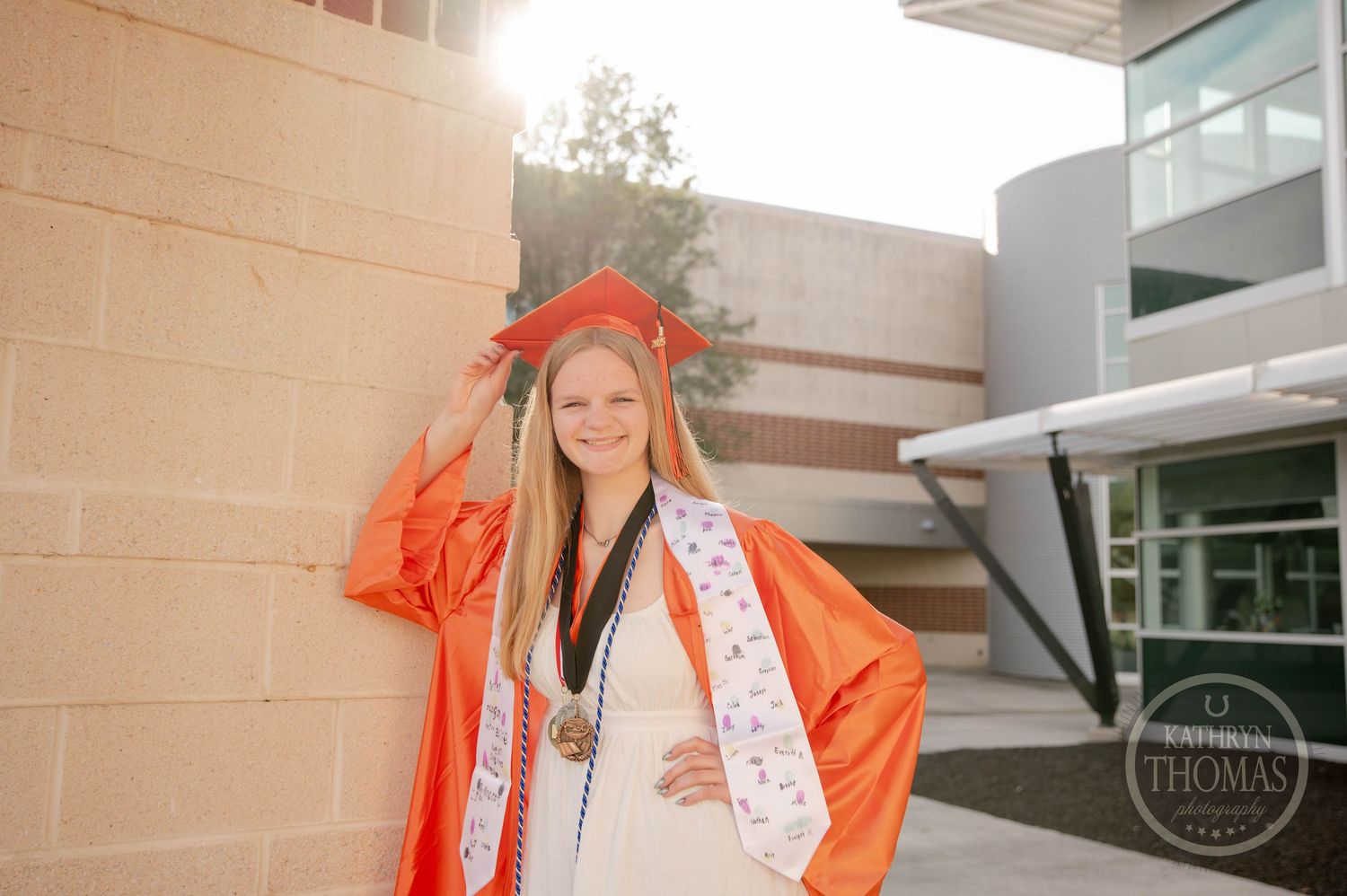 Graduate wearing bright orange academic regalia and decorative sash poses outdoors in sunlit campus setting.