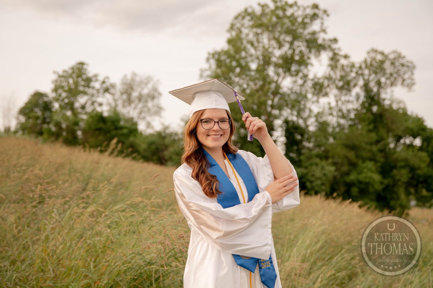 Graduates in white caps and gowns celebrate on a grassy hilltop during a summer graduation photoshoot.