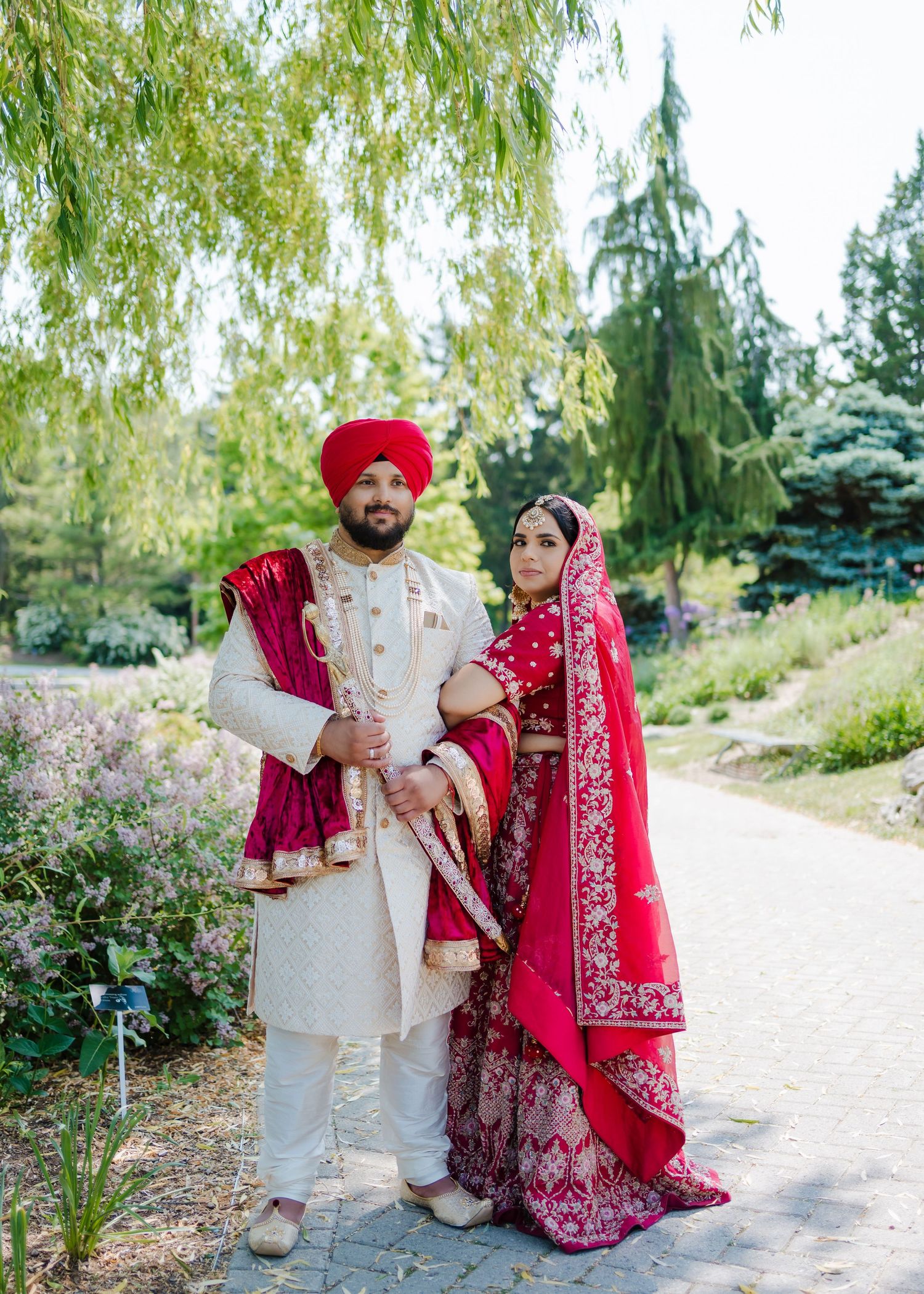 A couple in traditional Sikh wedding attire poses outdoors with willow trees and evergreens in the background.