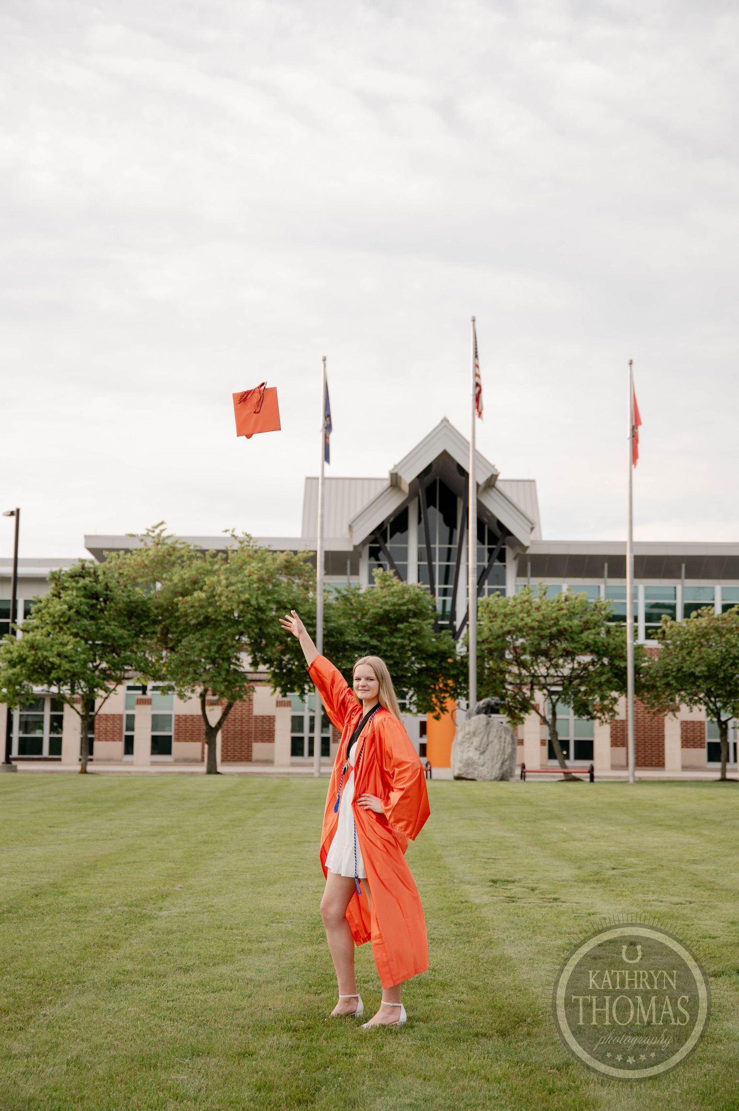 Graduate in orange cap and gown holds up red graduation cap with tassel in outdoor setting.