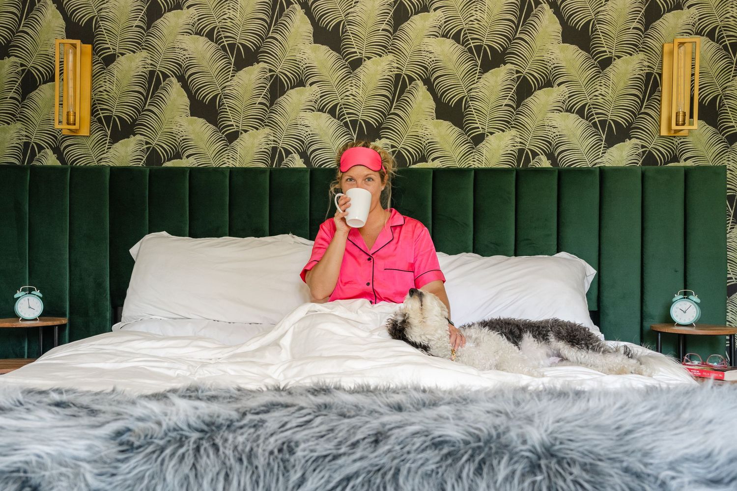Woman in pink pajamas and sleep mask sipping coffee on a luxurious bed with a dog and tropical wallpaper, in Orange County California.