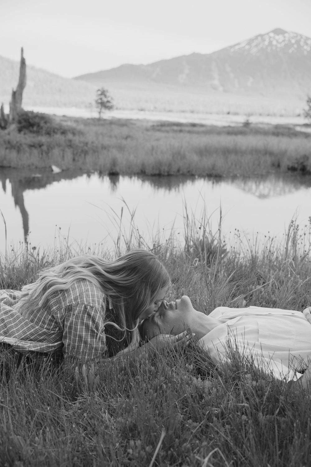 Black and white photo of romantic couple lying in grass near mountain lake, sharing an intimate moment.