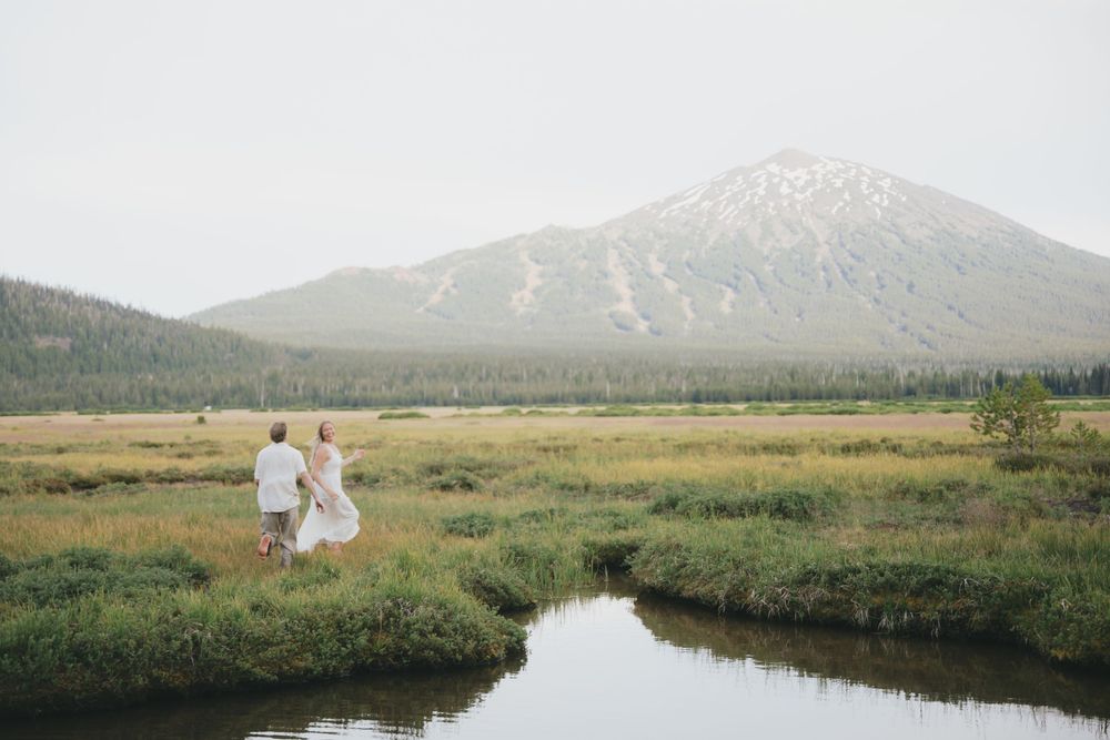 Couple walking hand-in-hand through a lush meadow with a stream and a snow-dusted mountain peak in the distance.