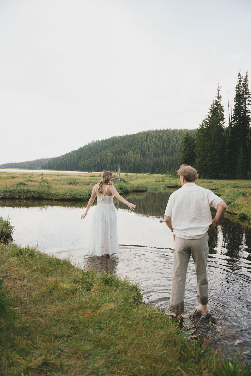 Bride and groom wading in a scenic mountain stream surrounded by evergreen trees and meadows.