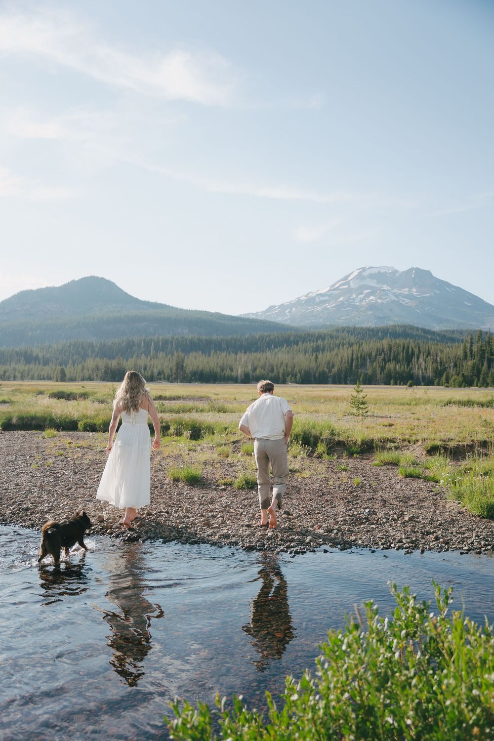 Couple and black dog walking on rocky riverbank with mountain and forest backdrop on a sunny day.