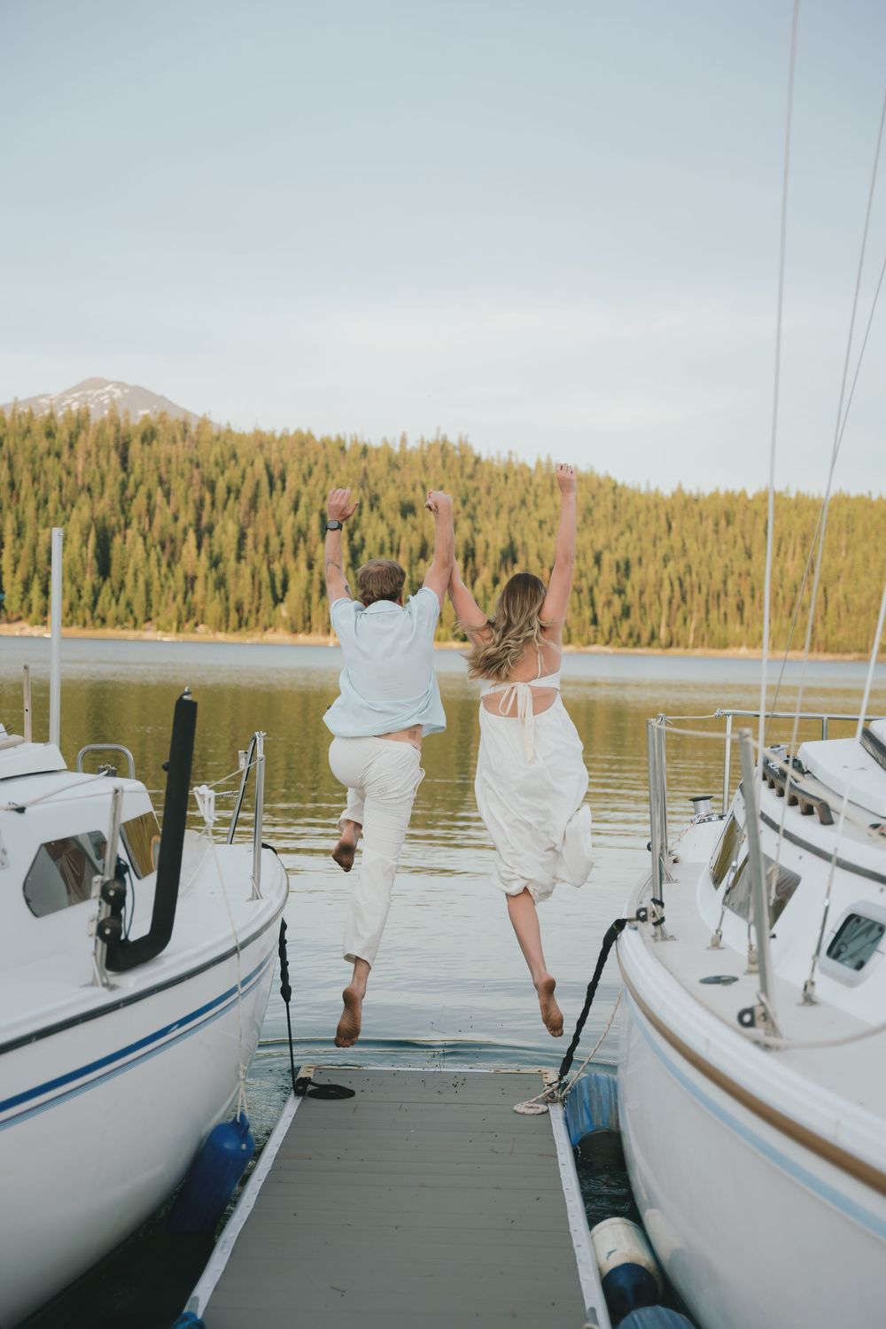 Joyful couple in white jumping on a boat dock surrounded by sailboats and scenic forest lake.