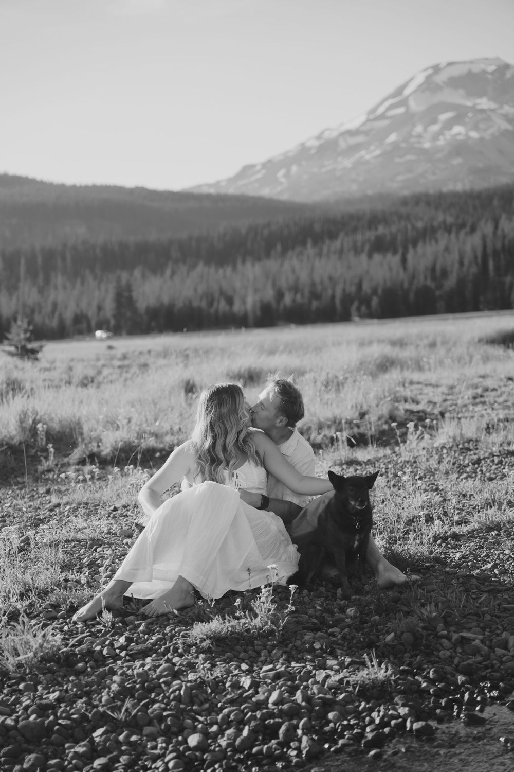 Black and white photo of a couple kissing outdoors with their dog, mountains and pine forest in the background.