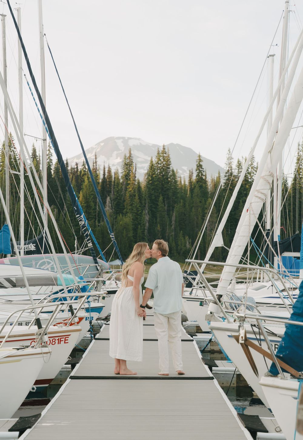 Couple kissing on a marina dock surrounded by sailboats with snow-capped mountains and pine trees in the background.