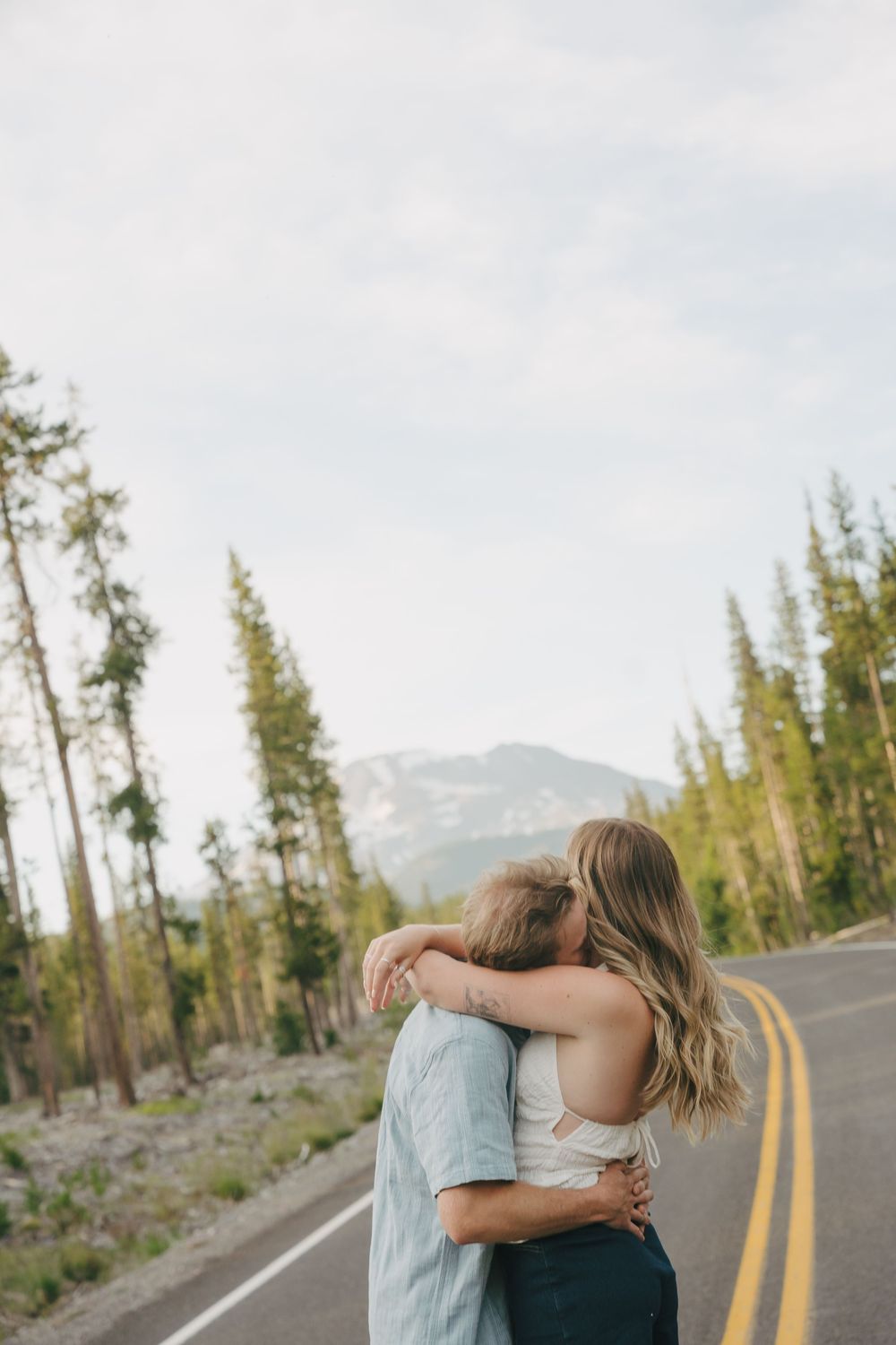 Couple embracing and kissing on a scenic mountain road surrounded by tall pine trees and misty peaks.