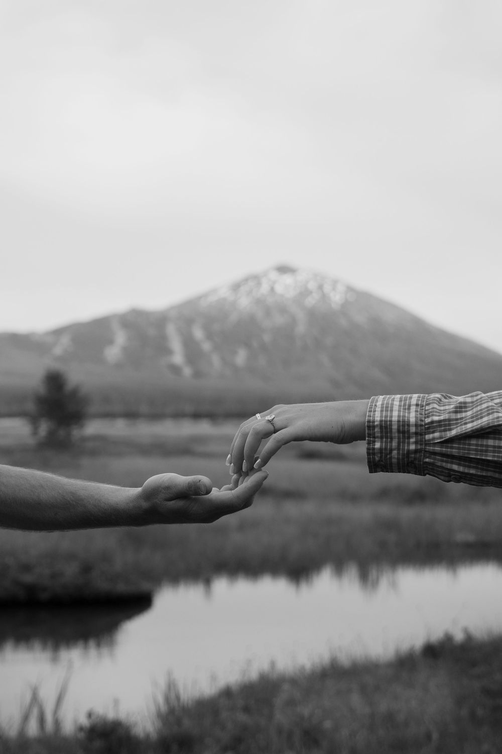 Black and white photo of two hands reaching toward each other with a mountain and reflective lake in the background.