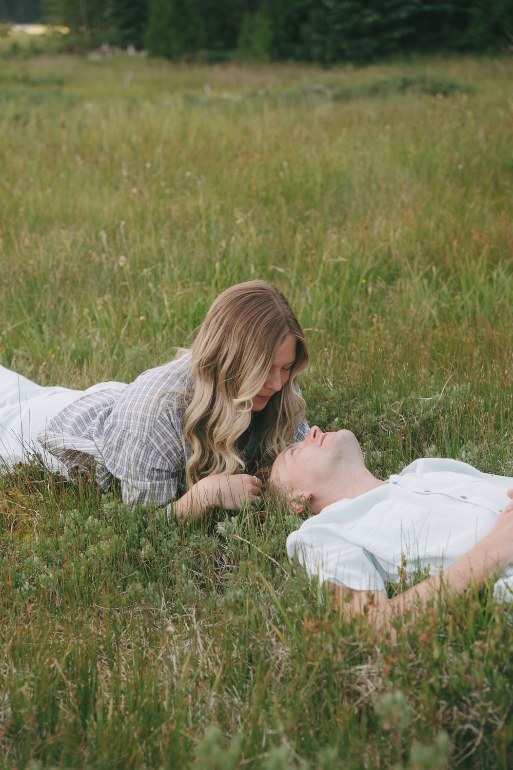 Couple lying in green meadow grass, woman with blonde hair leaning over man in romantic outdoor setting.
